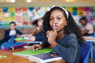 Buy stock photo Shot of a young girl sitting in her school classroom with her finger on her lips in a hushing gesture