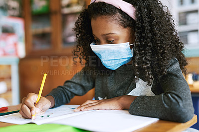 Buy stock photo Shot of a young girl sitting in her classroom at school and writing in her workbook while wearing a face mask