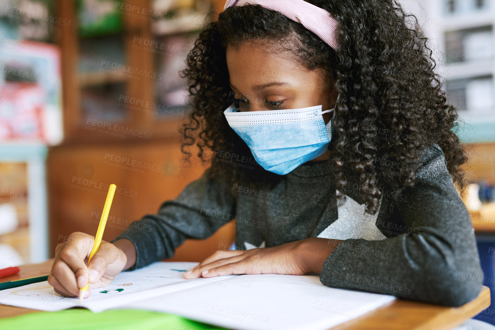 Buy stock photo Shot of a young girl sitting in her classroom at school and writing in her workbook while wearing a face mask