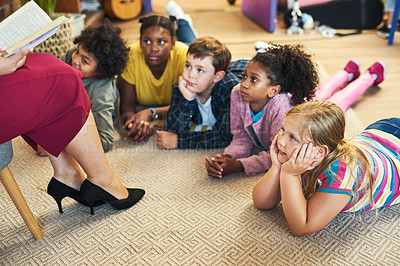 Buy stock photo Shot of a diverse group of children lying on their classroom floor and listening to their teacher read a book