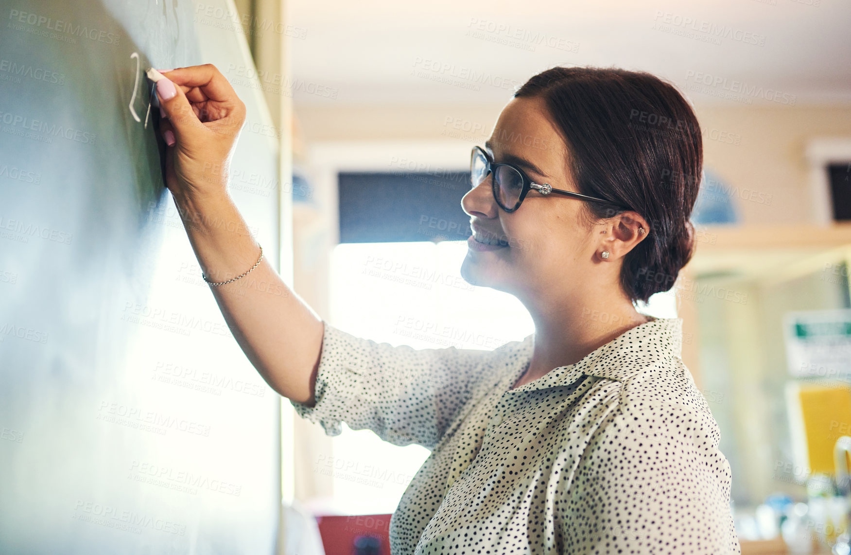 Buy stock photo Shot of an attractive young teacher standing alone and writing on the blackboard during a lesson in her classroom