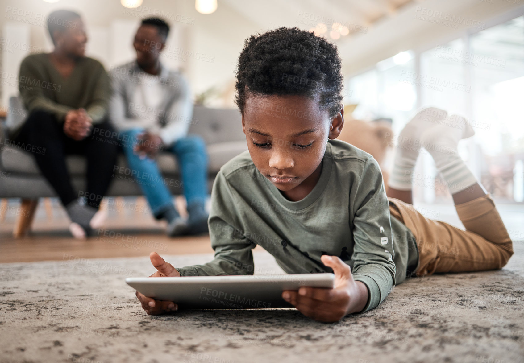 Buy stock photo Shot of an adorable little boy using a digital tablet with his family in the background at home