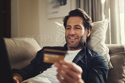 Buy stock photo Shot of a handsome young man using his laptop and credit card while relaxing on a couch at home