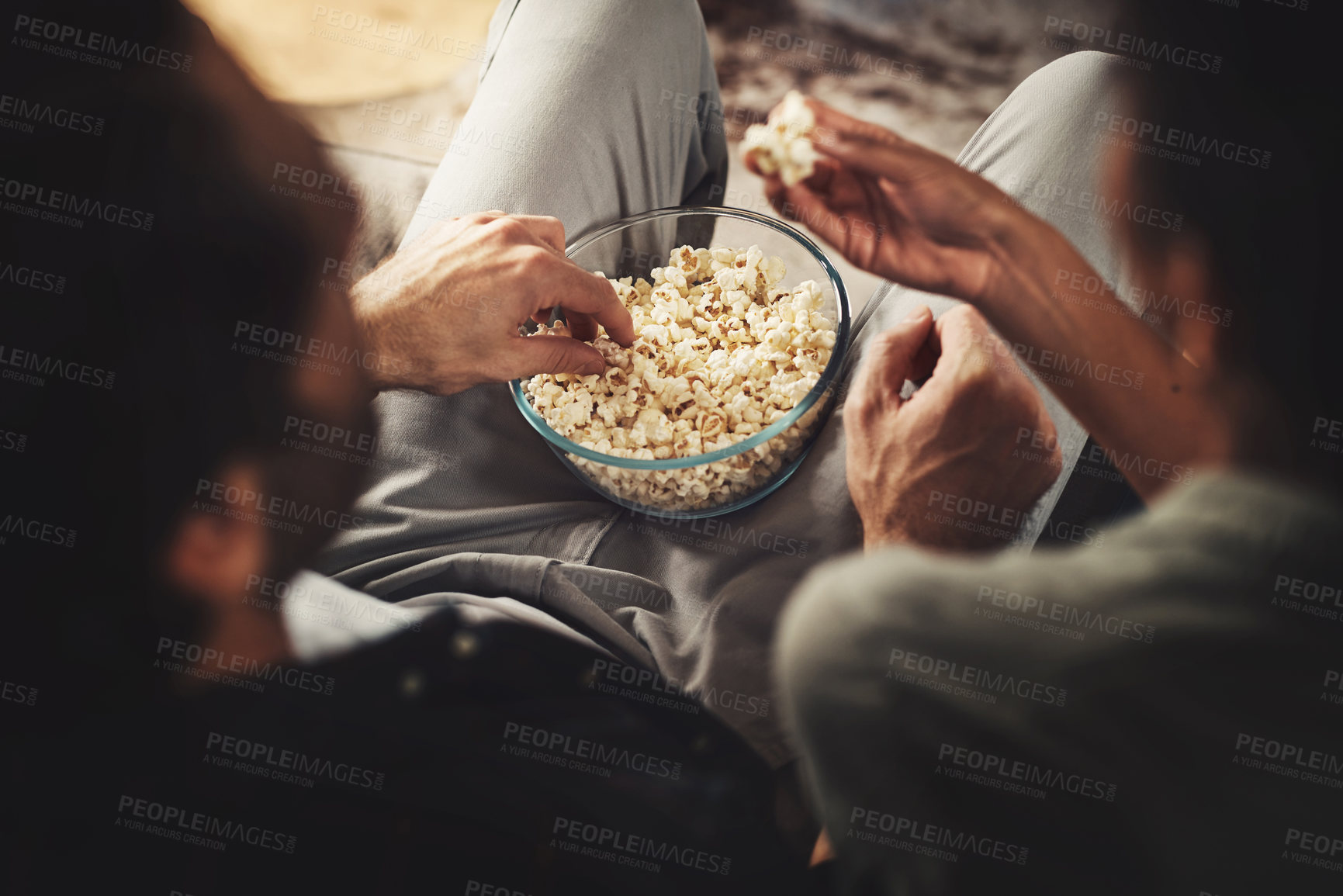 Buy stock photo Rearview shot of a young couple sitting on sofa snacking on popcorn together at home
