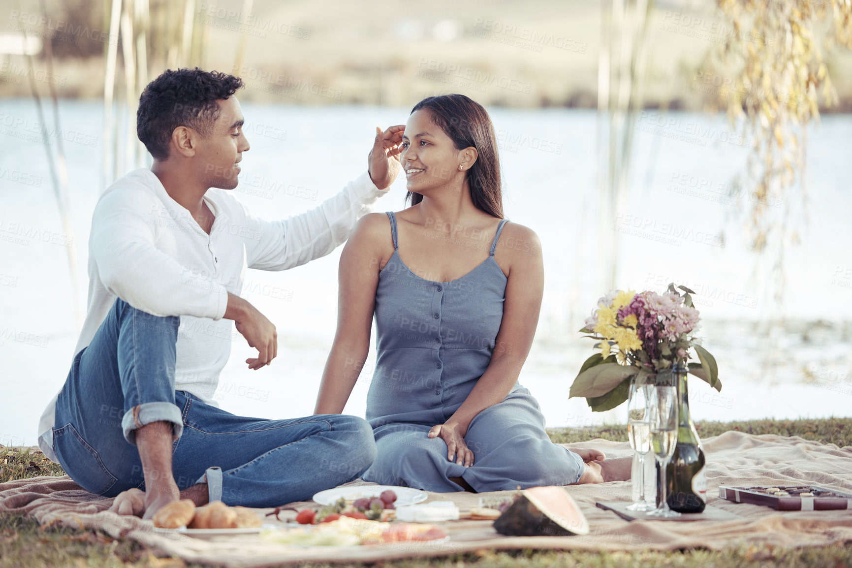 Buy stock photo Full length shot of a young couple sitting by a lake on a date and enjoying a picnic