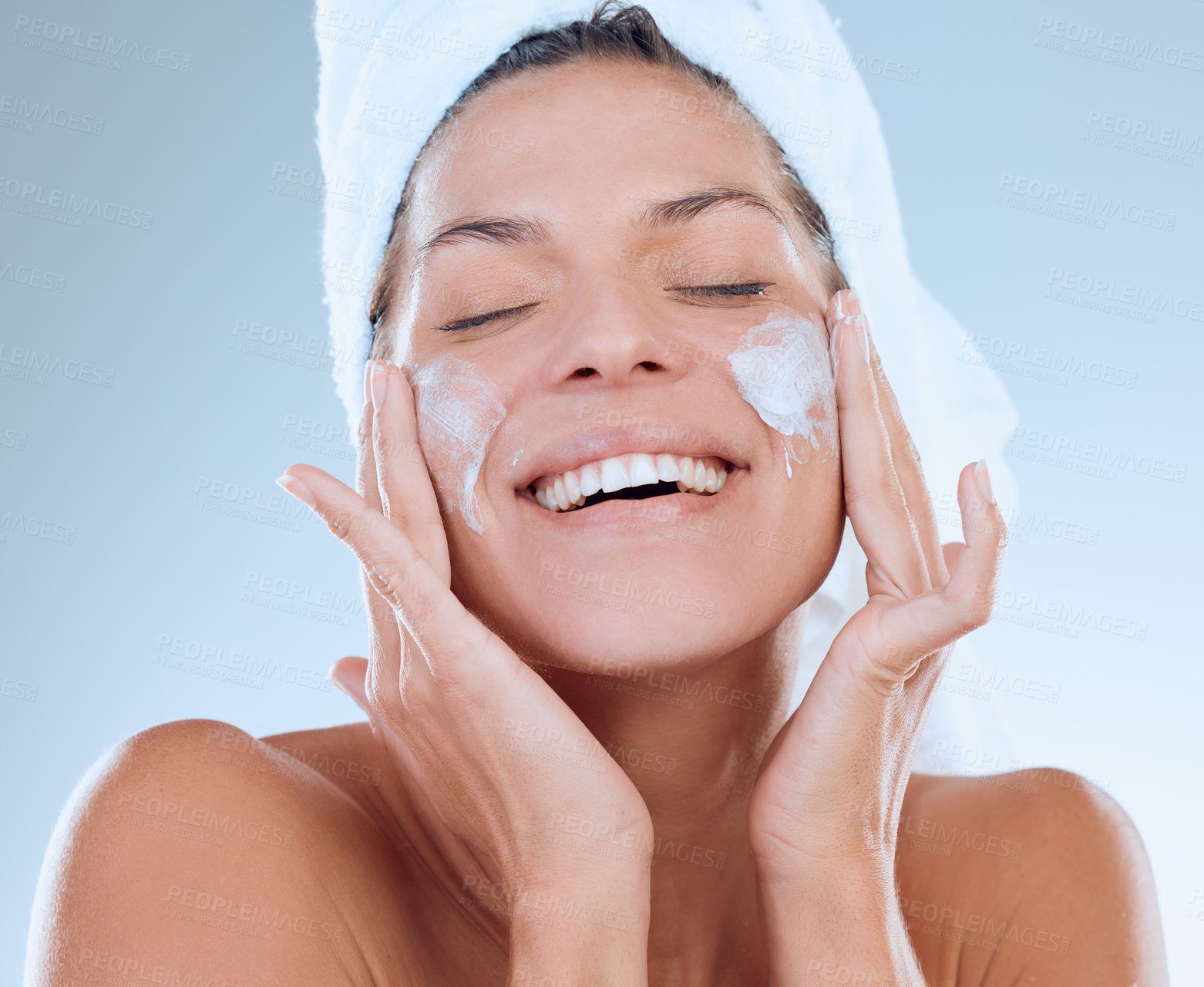 Buy stock photo Studio shot of a young woman applying moisturiser to her face after a shower against a blue background