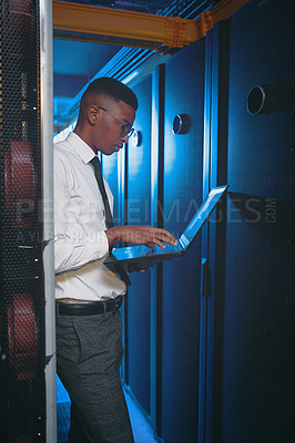 Buy stock photo Shot of a young IT specialist standing alone in the server room and using his laptop