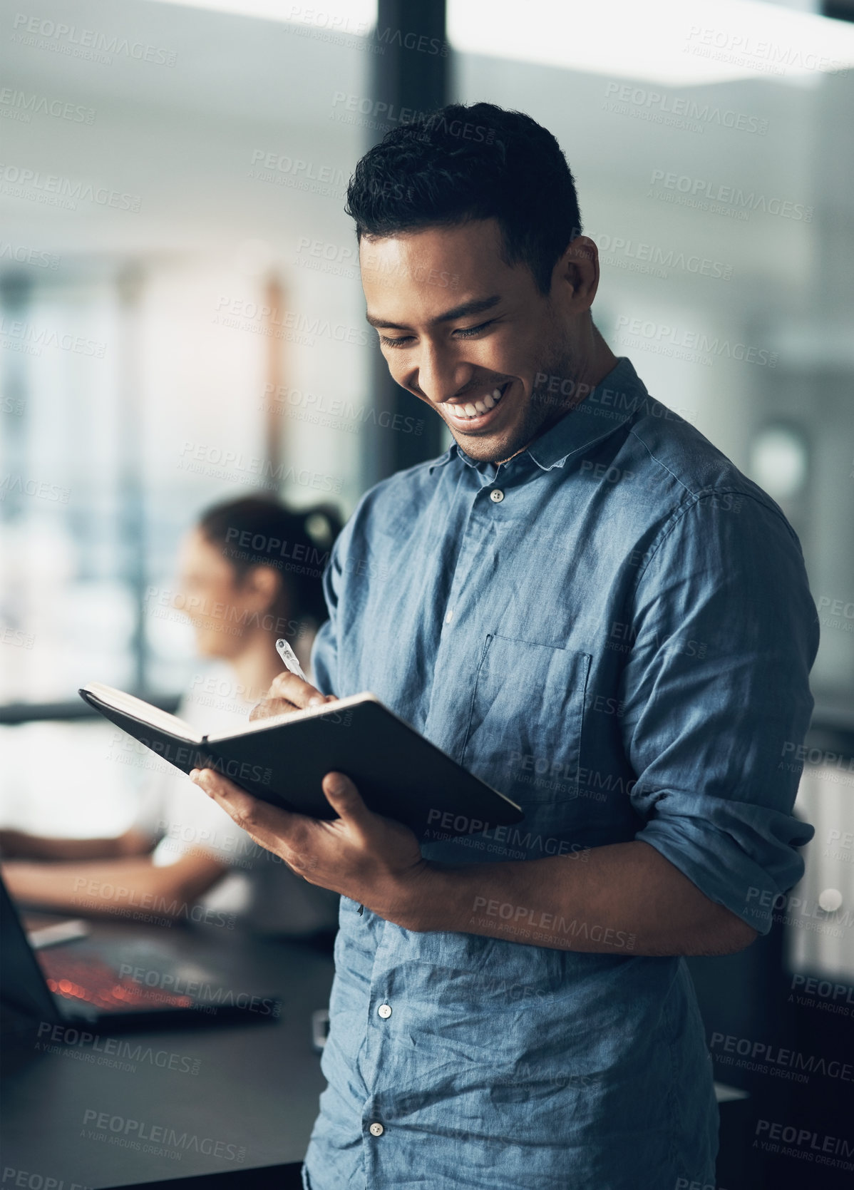 Buy stock photo Shot of a happy young man making notes in a book in a modern office