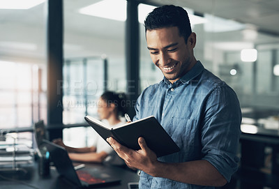 Buy stock photo Shot of a happy young man making notes in a book in a modern office