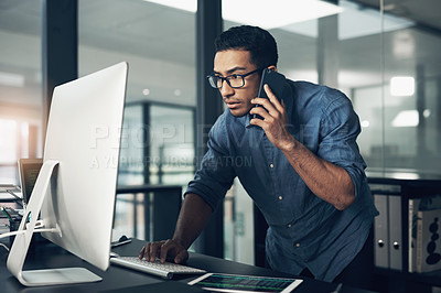 Buy stock photo Shot of a young man using his cellphone while working on a computer in a modern office