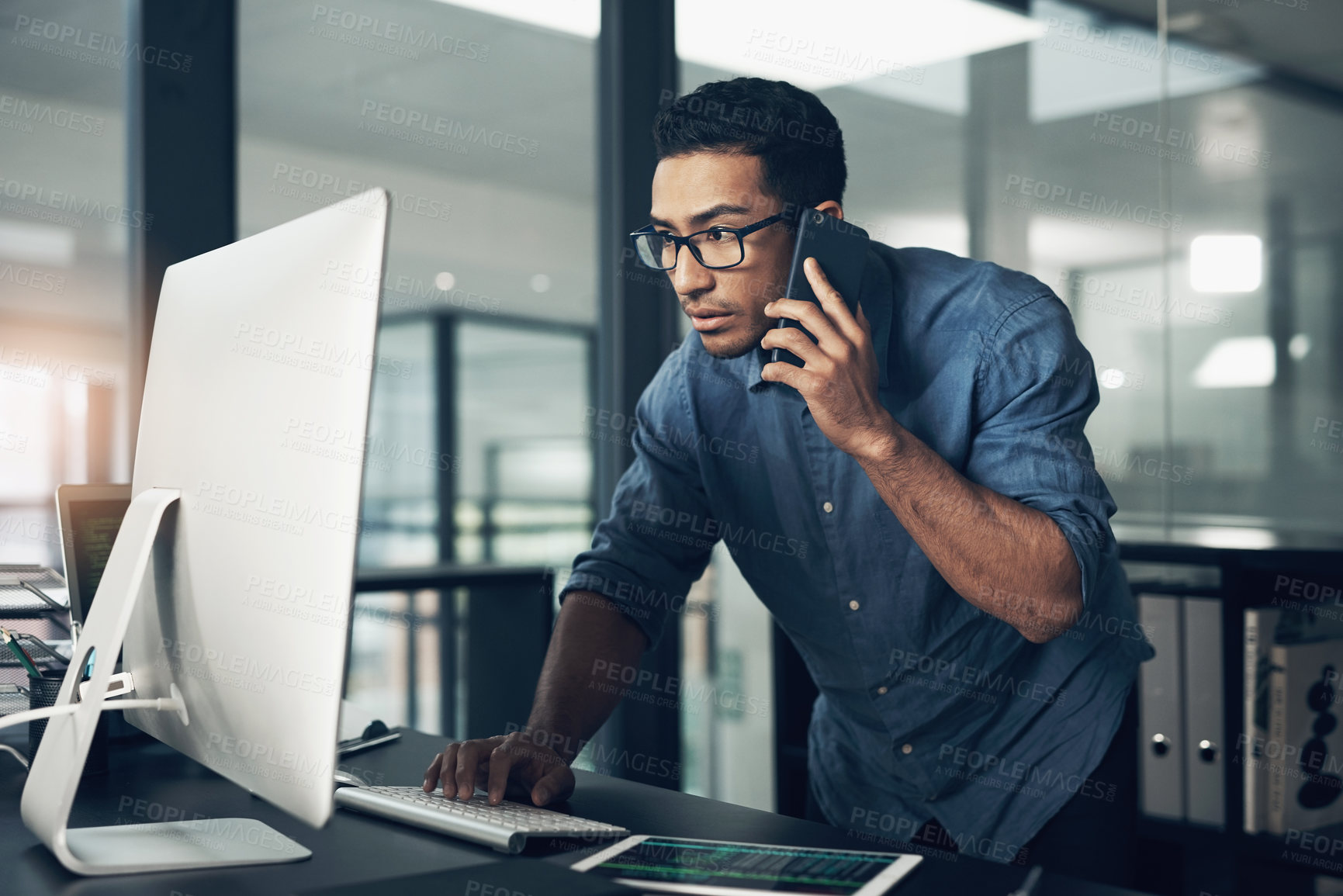 Buy stock photo Shot of a young man using his cellphone while working on a computer in a modern office