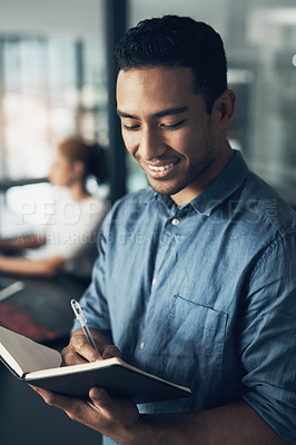 Buy stock photo Shot of a happy young man making notes in a book in a modern office