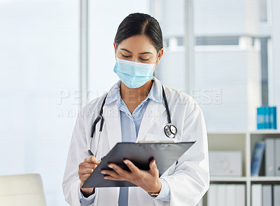 Buy stock photo Shot of a doctor wearing a face mask and standing alone while writing on her clipboard in her clinic