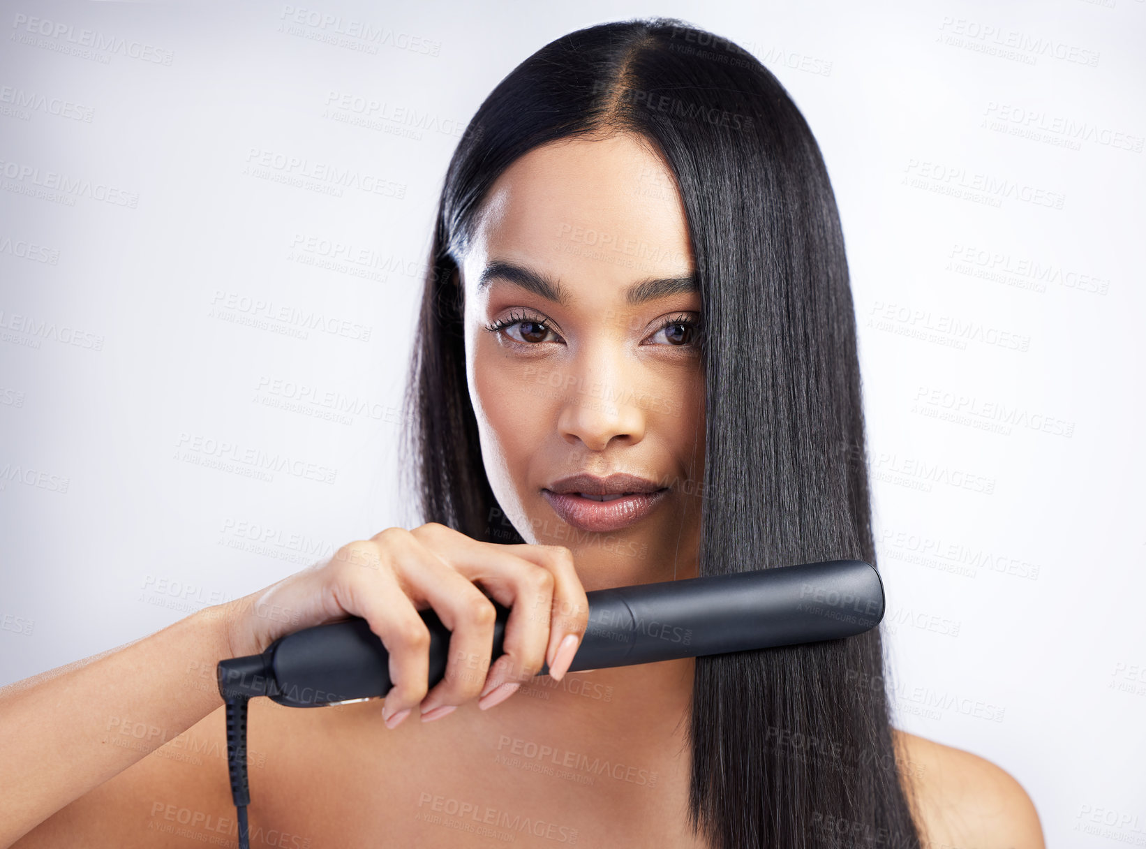 Buy stock photo Shot of an attractive young woman standing alone in the studio and styling her hair with a hair straightener