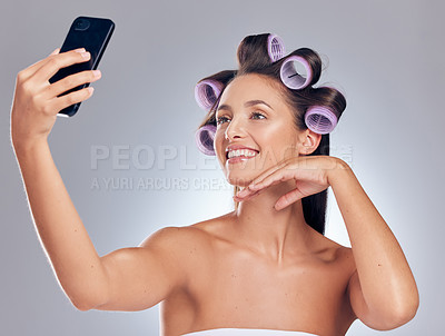 Buy stock photo Shot of a young woman standing in the studio with rollers in her hair and taking selfies with her cellphone