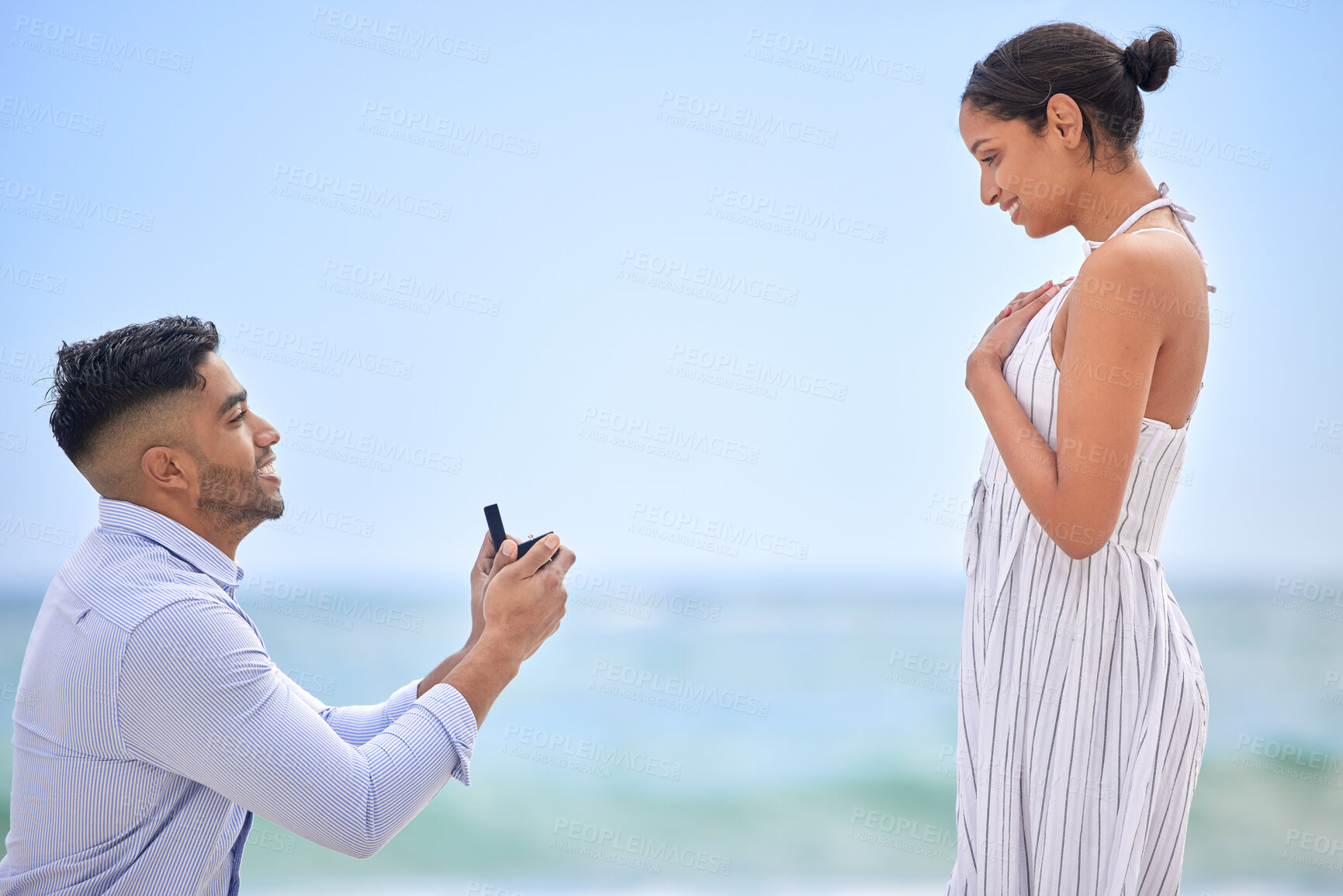 Buy stock photo Engagement, proposal and romance with a couple on the beach for a milestone on a blue sky background. Love, summer or nature with a man down on one knee to ask his girlfriend the marriage question