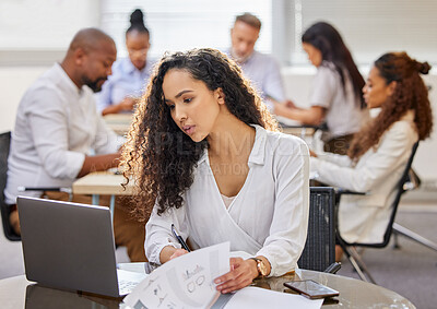 Buy stock photo Shot of a young businesswoman going through paperwork while working on a laptop in an office with her colleagues in the background