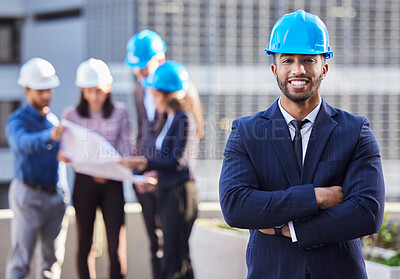 Buy stock photo Shot of a young businessman standing with his arms folded and wearing a hardhat while his colleagues stand behind him