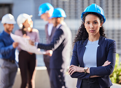 Buy stock photo Shot of a young businesswoman standing with her arms folded and wearing a hardhat while her colleagues stand behind her