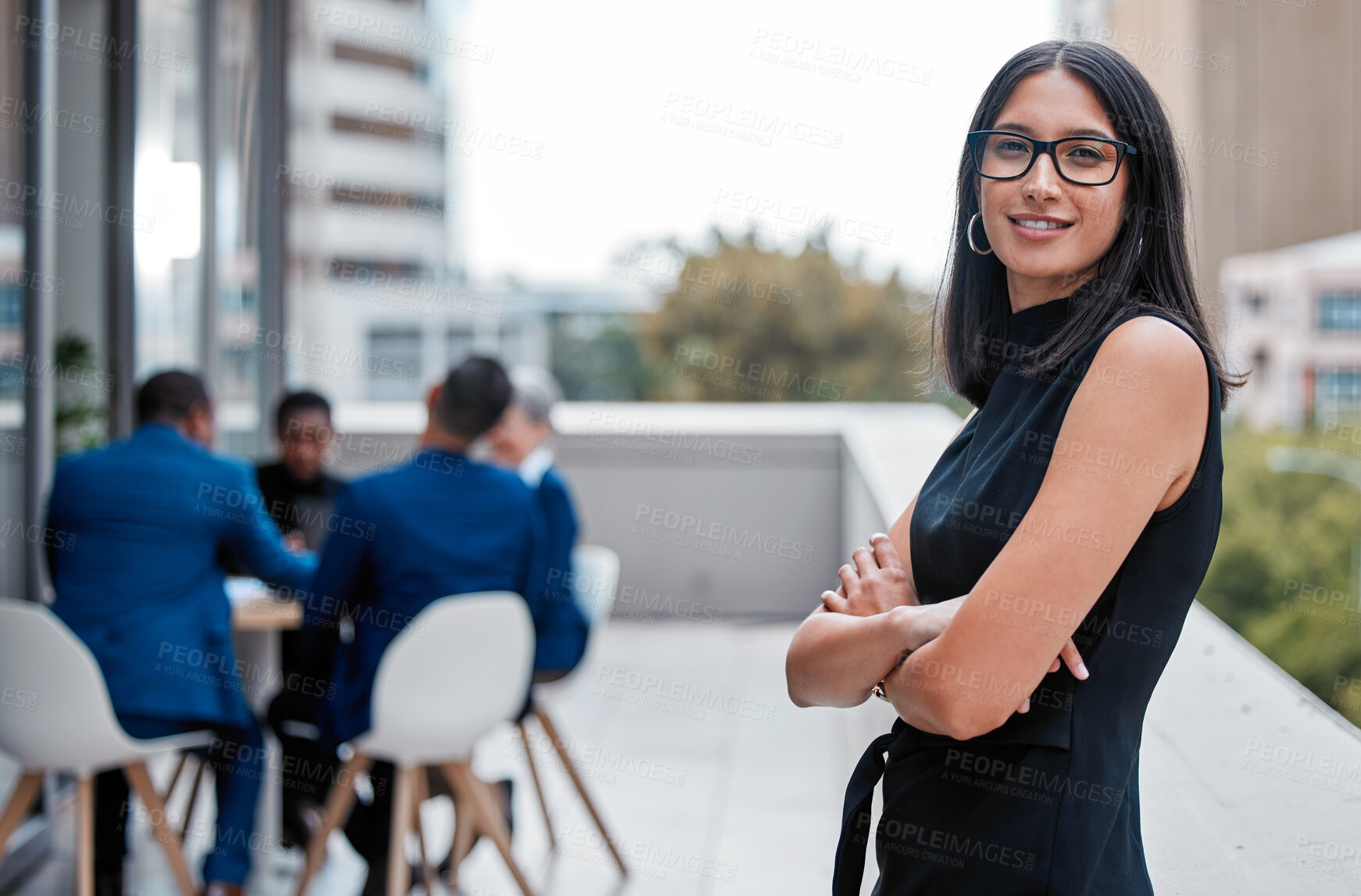 Buy stock photo Cropped portrait of an attractive young businesswoman standing with her arms folded outside on the office balcony with her colleagues in the background