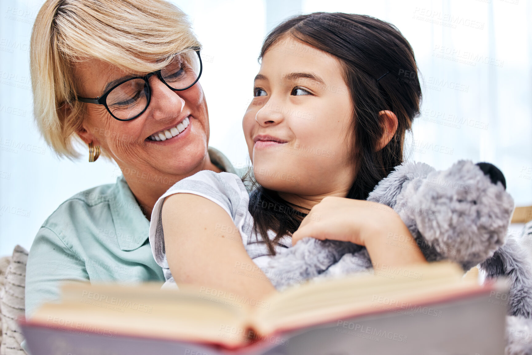 Buy stock photo Happy, book and senior woman with child reading for knowledge with teddy bear at home together. Smile, bonding and elderly grandmother relaxing and enjoying story with girl kid holding toy at house.