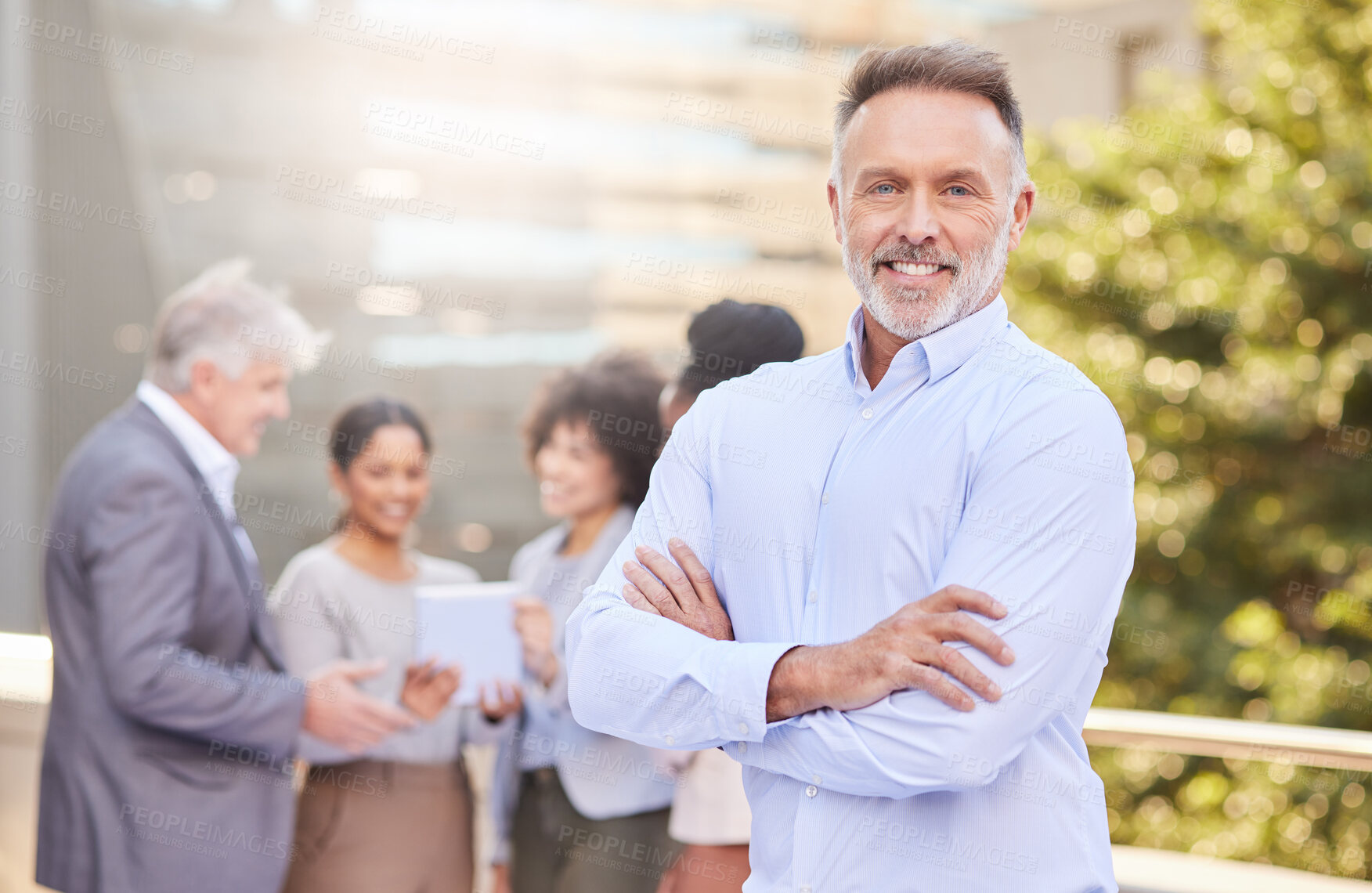 Buy stock photo Shot of a handsome mature businessman standing outside with his arms folded while his colleagues discuss behind him