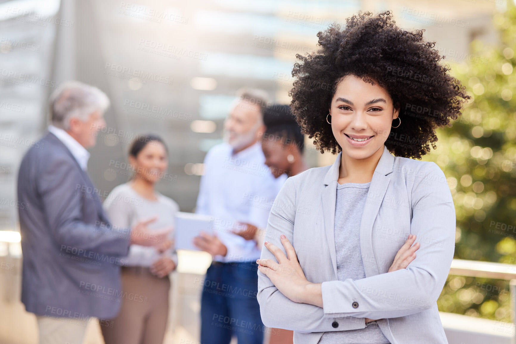 Buy stock photo Shot of an attractive young businesswoman standing outside with her arms folded while her colleagues discuss behind her