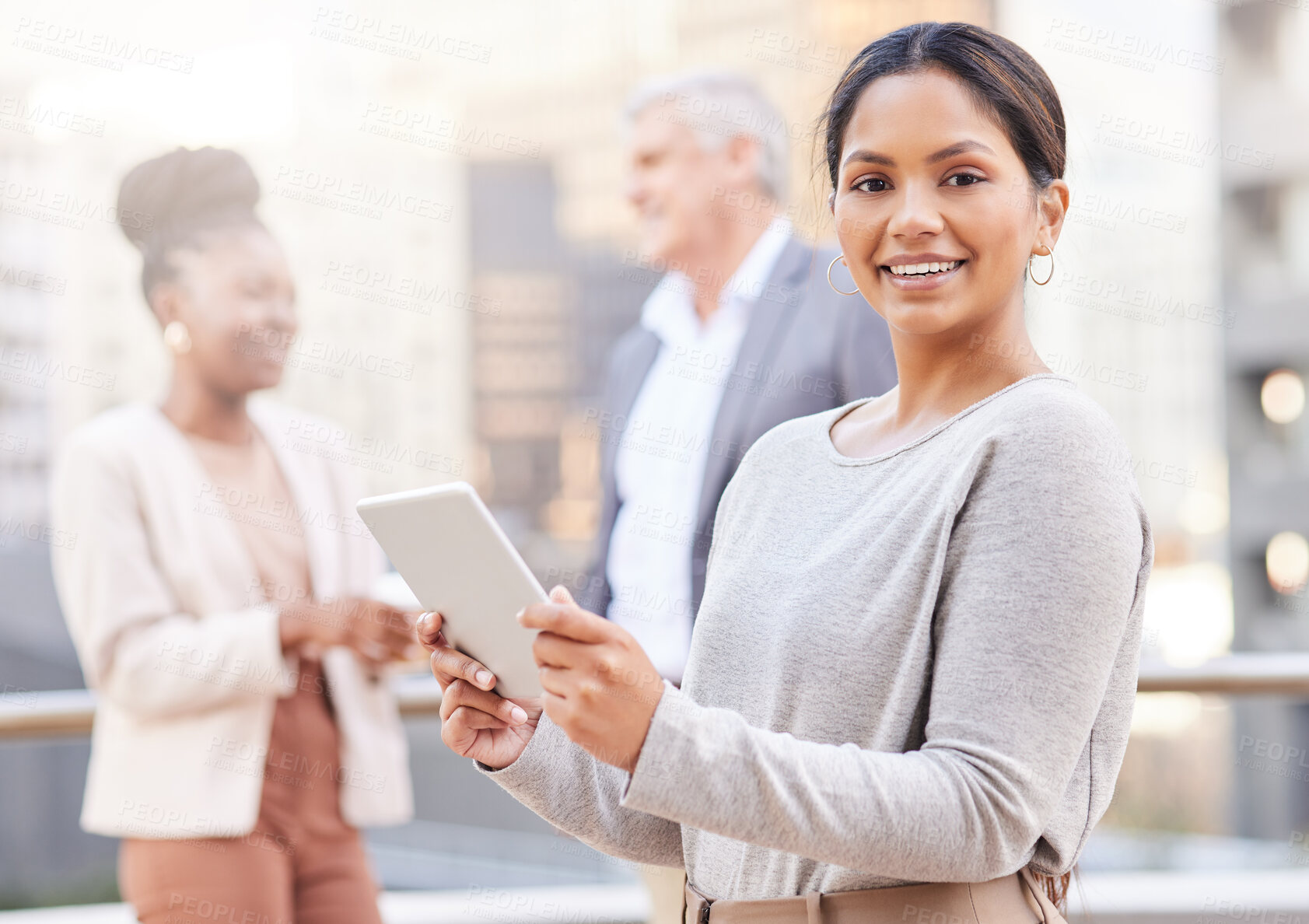 Buy stock photo Shot of an attractive young businesswoman standing outside and using a digital tablet while her colleagues discuss behind her