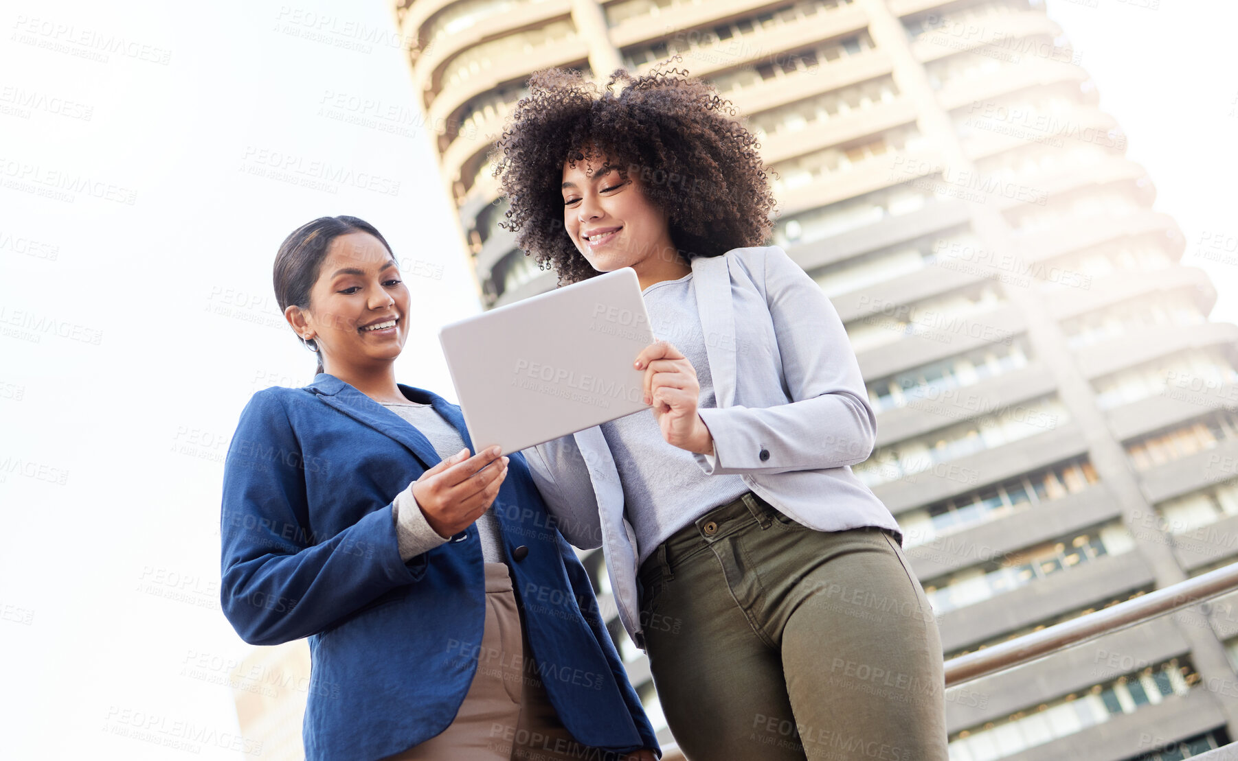 Buy stock photo Low angle shot of two young businesswomen standing outside on the balcony and using a digital tablet