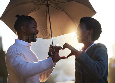 Buy stock photo Shot of a young man and woman making a heart shape with their hands while standing under an umbrella together on a rainy day in the city