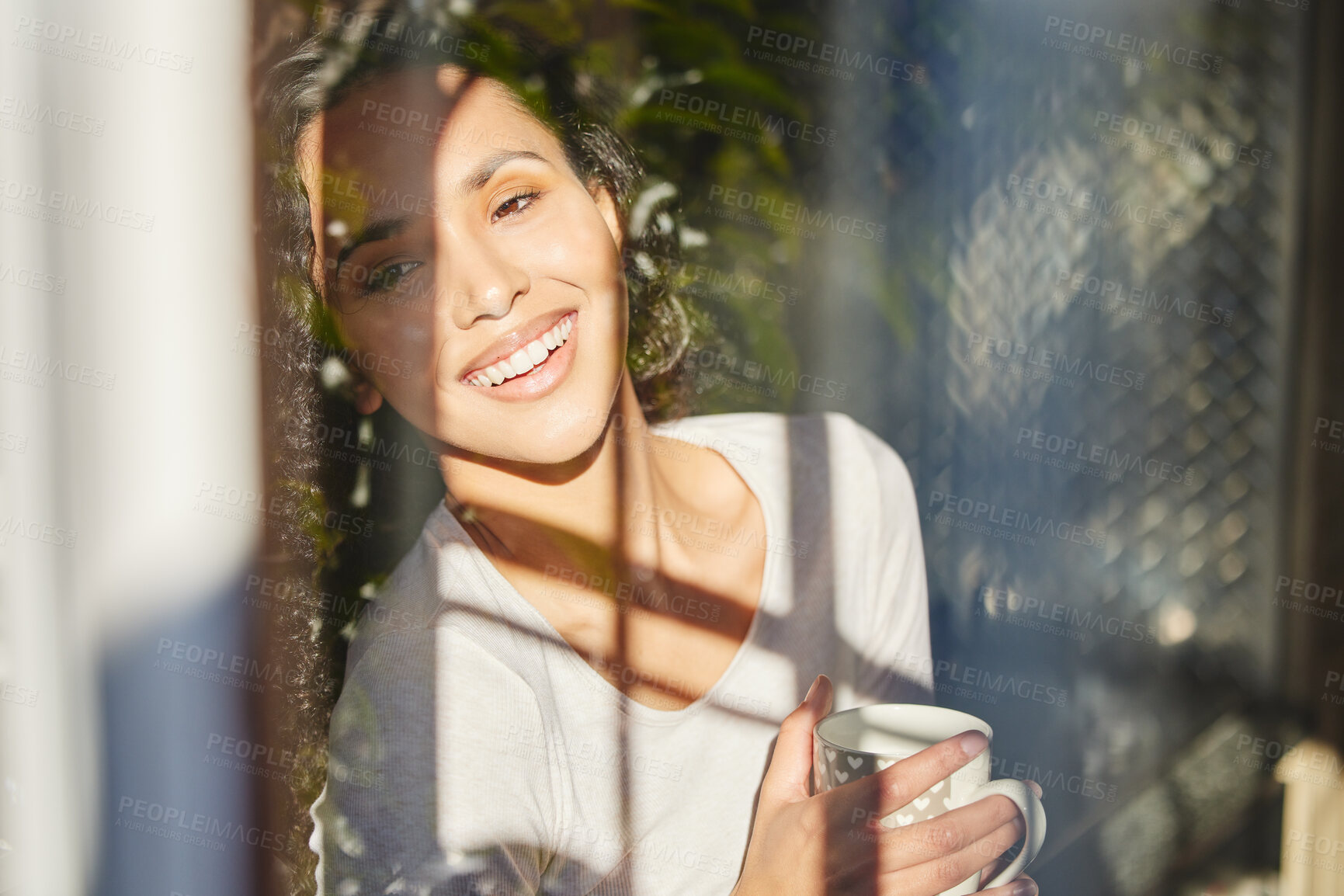 Buy stock photo Shot of a young woman standing and enjoying a cup of coffee while looking out of her window