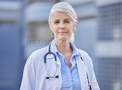 Buy stock photo Shot of a mature female doctor standing with her arms crossed against a city background