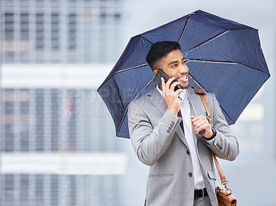 Buy stock photo Shot of a young businessman holding an umbrella while talking on a cellphone on a rainy day in the city