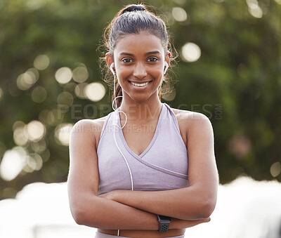 Buy stock photo Portrait of a sporty young woman wearing earphones and standing with her arms crossed while exercising outdoors