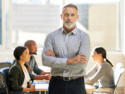 Buy stock photo Cropped portrait of a handsome mature businessman standing with his arms crossed in the boardroom while a meeting takes place behind him