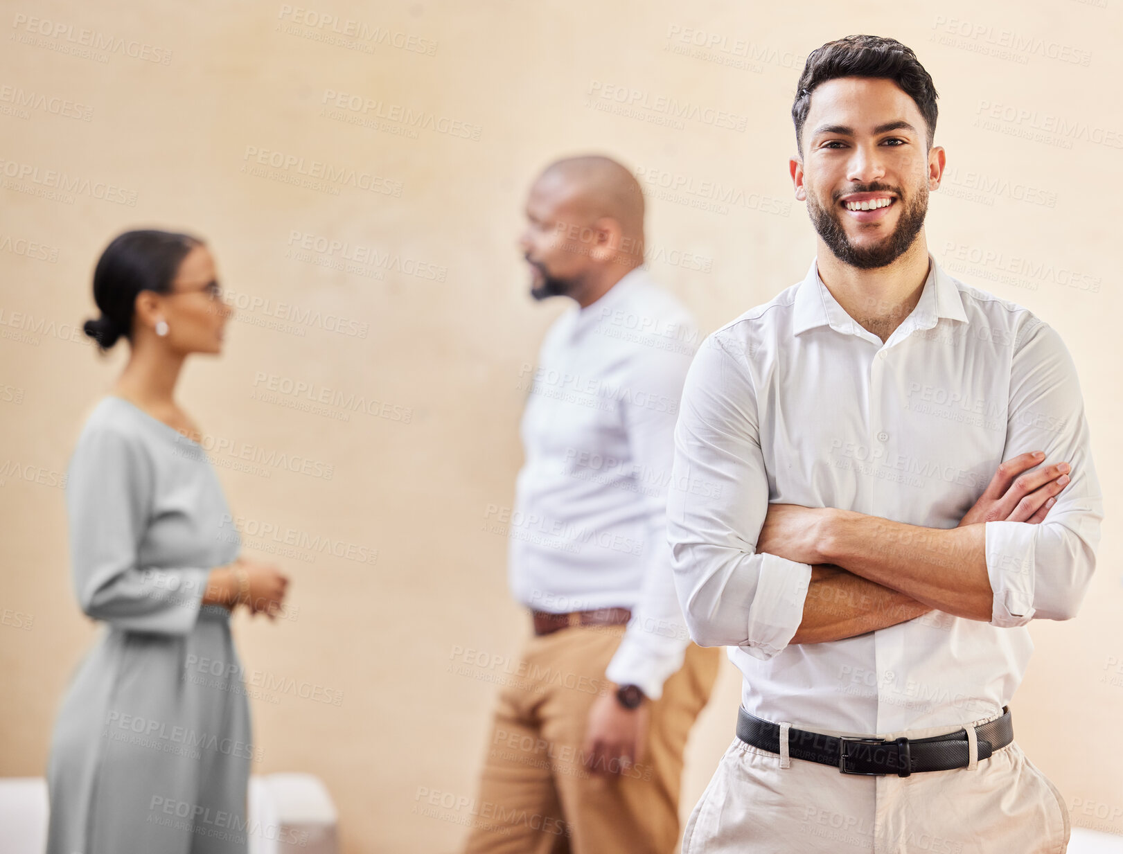 Buy stock photo Cropped portrait of a handsome young businessman standing with his arms crossed in the office with colleagues in the background