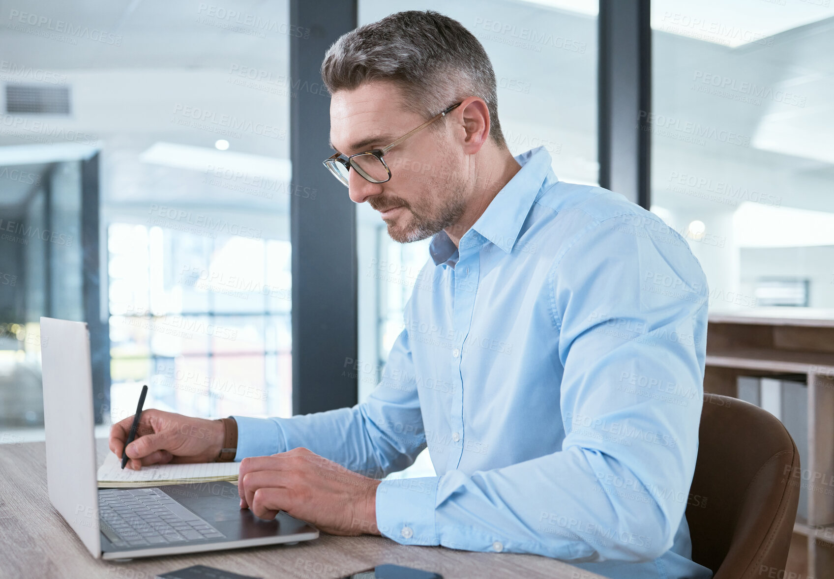 Buy stock photo Shot of a mature businessman writing notes while working on a laptop in an office