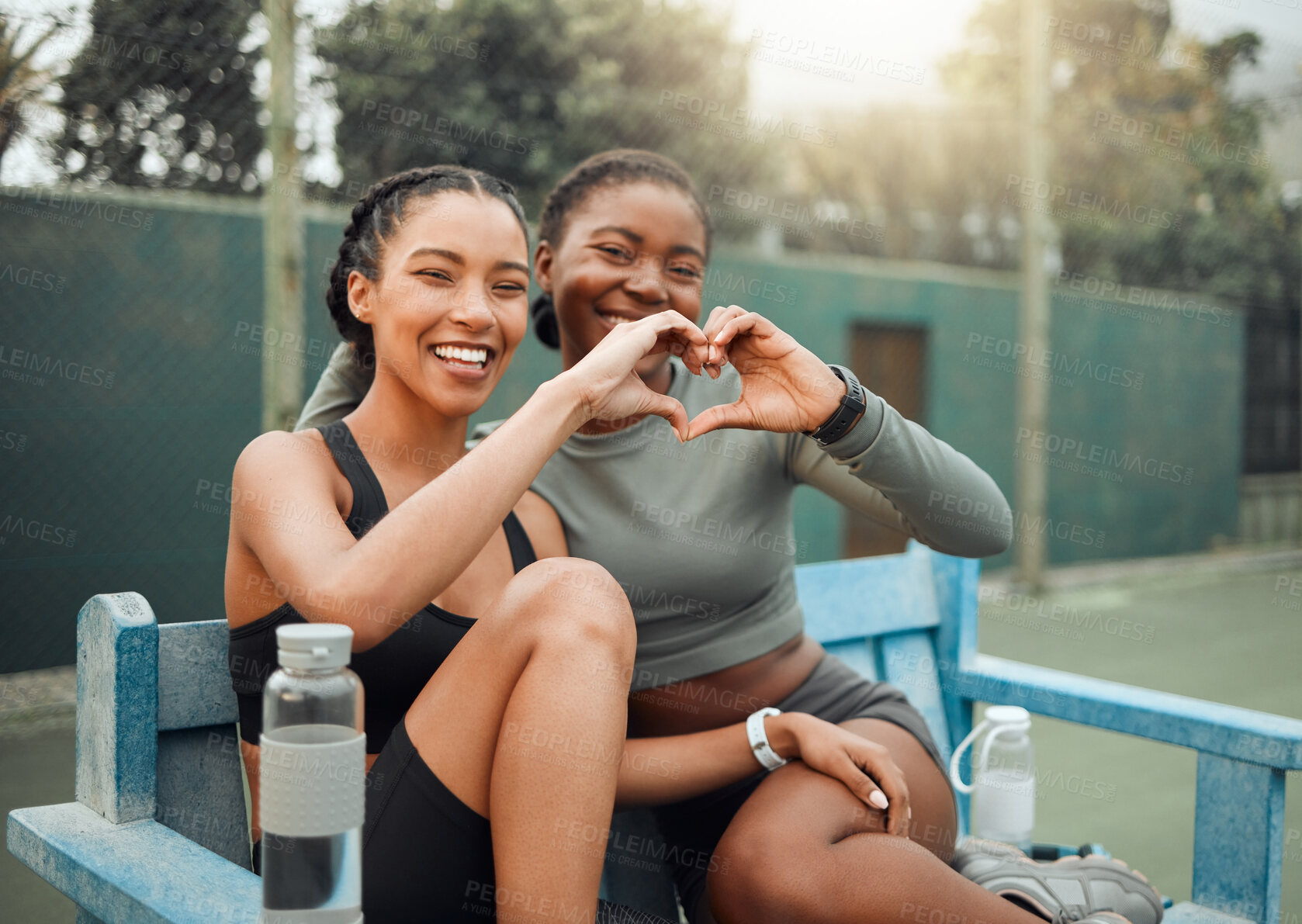 Buy stock photo Cropped portrait of two attractive young female athletes making a heart shape with their hands while sitting on a bench outside during a workout