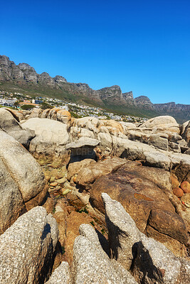 Buy stock photo Coastal rocks with view of the mountains and city on blue sky background with copy space. Bright summer landscape of big nature stones with rough texture details near luxury houses in Cape Town