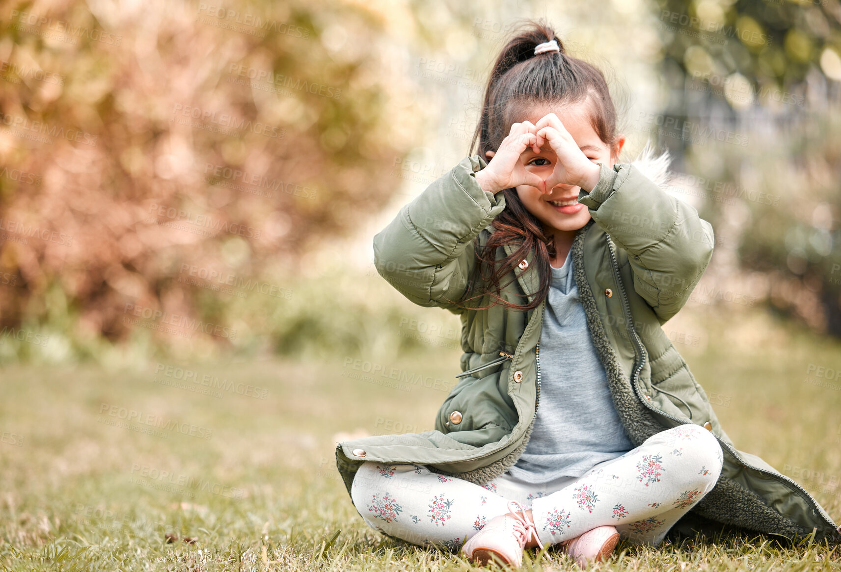 Buy stock photo Shot of a little girl forming a heart shape over her eye while sitting outside