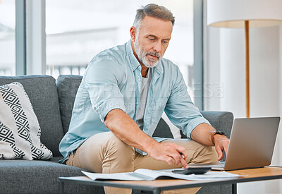 Buy stock photo Shot of a mature man sitting alone on his sofa and using his laptop while calculating his finances
