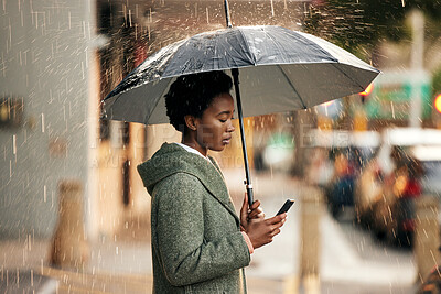 Buy stock photo Shot of a young businesswoman using an umbrella and a smartphone while going for a walk in the rain against an urban background