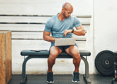 Buy stock photo Shot of a muscular young man checking his watch while using a laptop in a gym