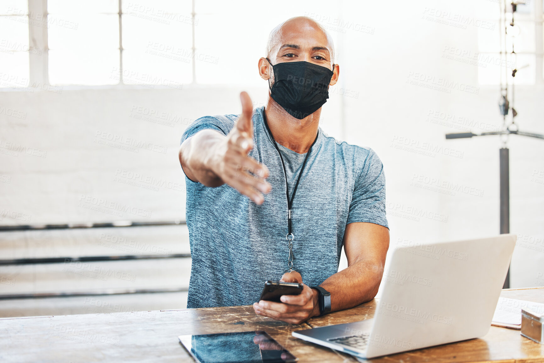 Buy stock photo Portrait of a muscular young man wearing a face mask and extending a handshake while using a laptop in a gym