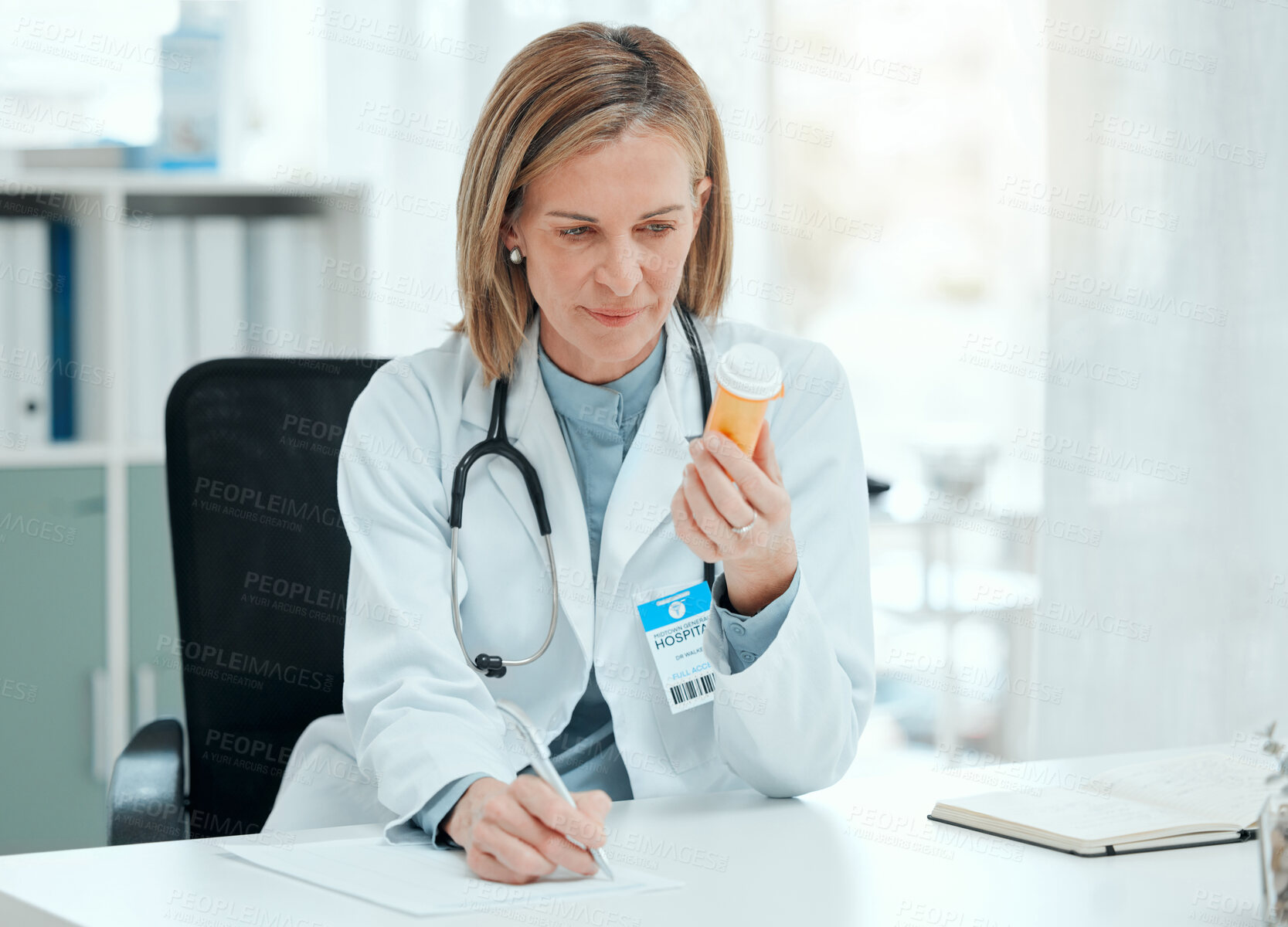 Buy stock photo Shot of a mature doctor sitting alone in her clinic and writing notes while holding a bottle of pills