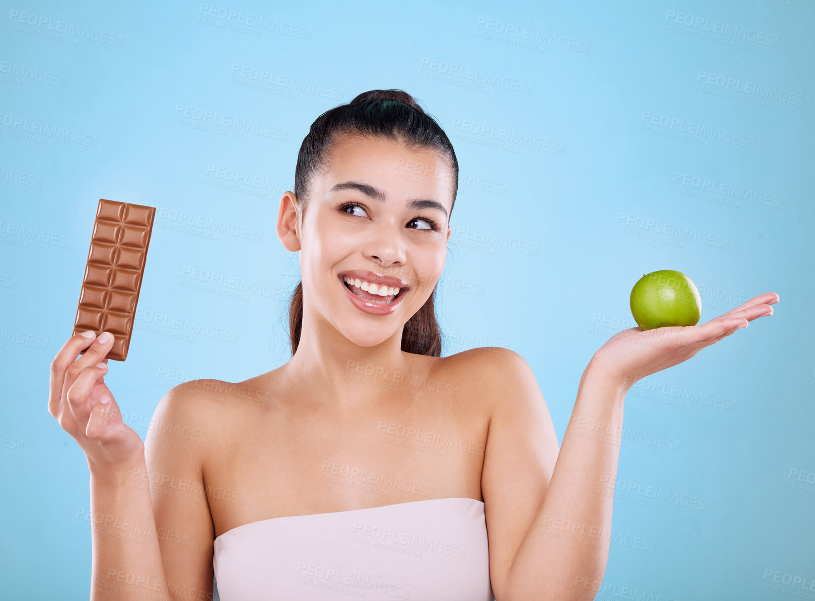 Buy stock photo Studio shot an attractive young woman deciding between a chocolate and an apple against a blue background