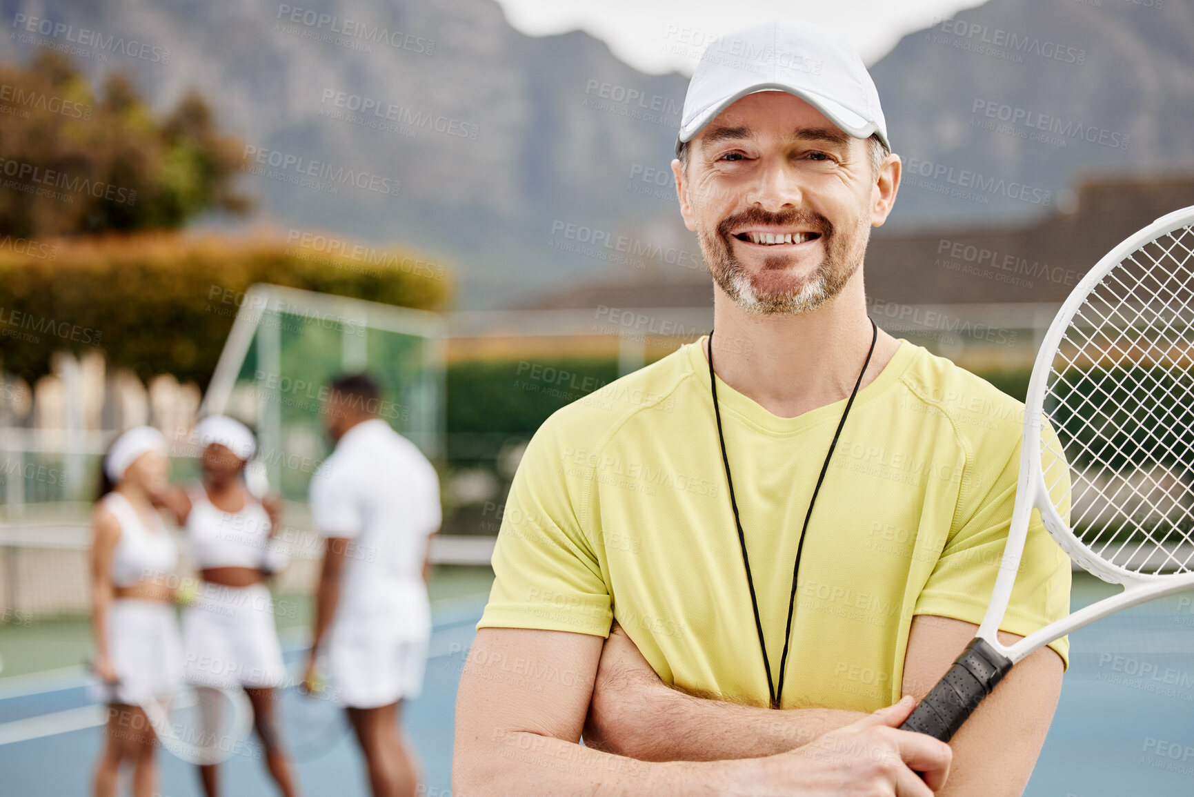 Buy stock photo Cropped portrait of a handsome mature male tennis coach standing outside with his arms folded and his students in the background