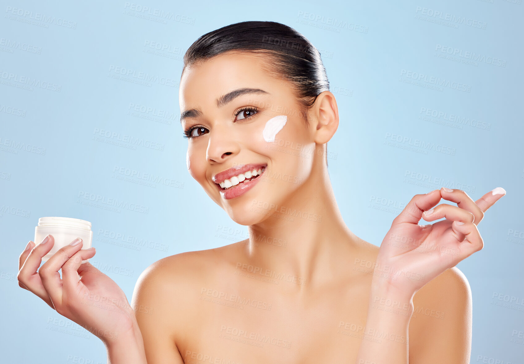 Buy stock photo Studio portrait of an attractive young woman applying moisturiser on her face against a blue background