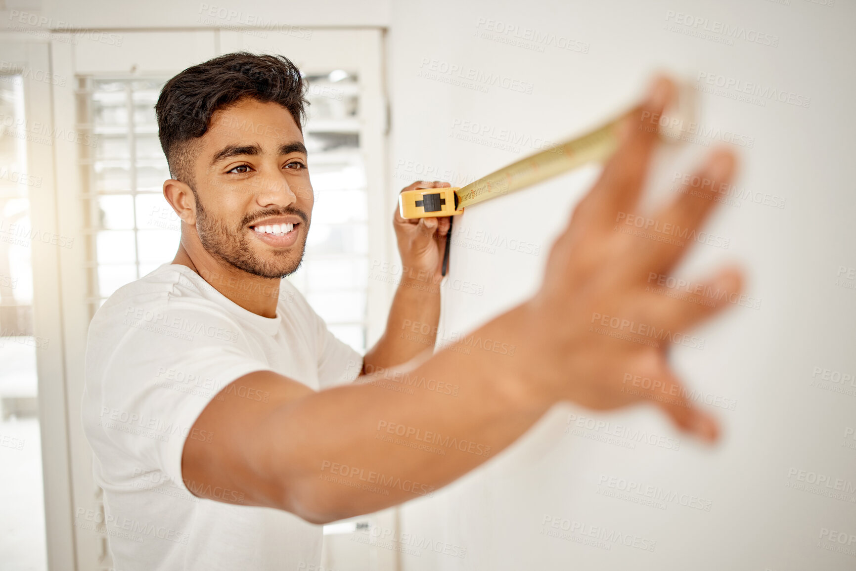 Buy stock photo Shot of a handsome young man standing alone in his home and using measuring tape to measure a wall