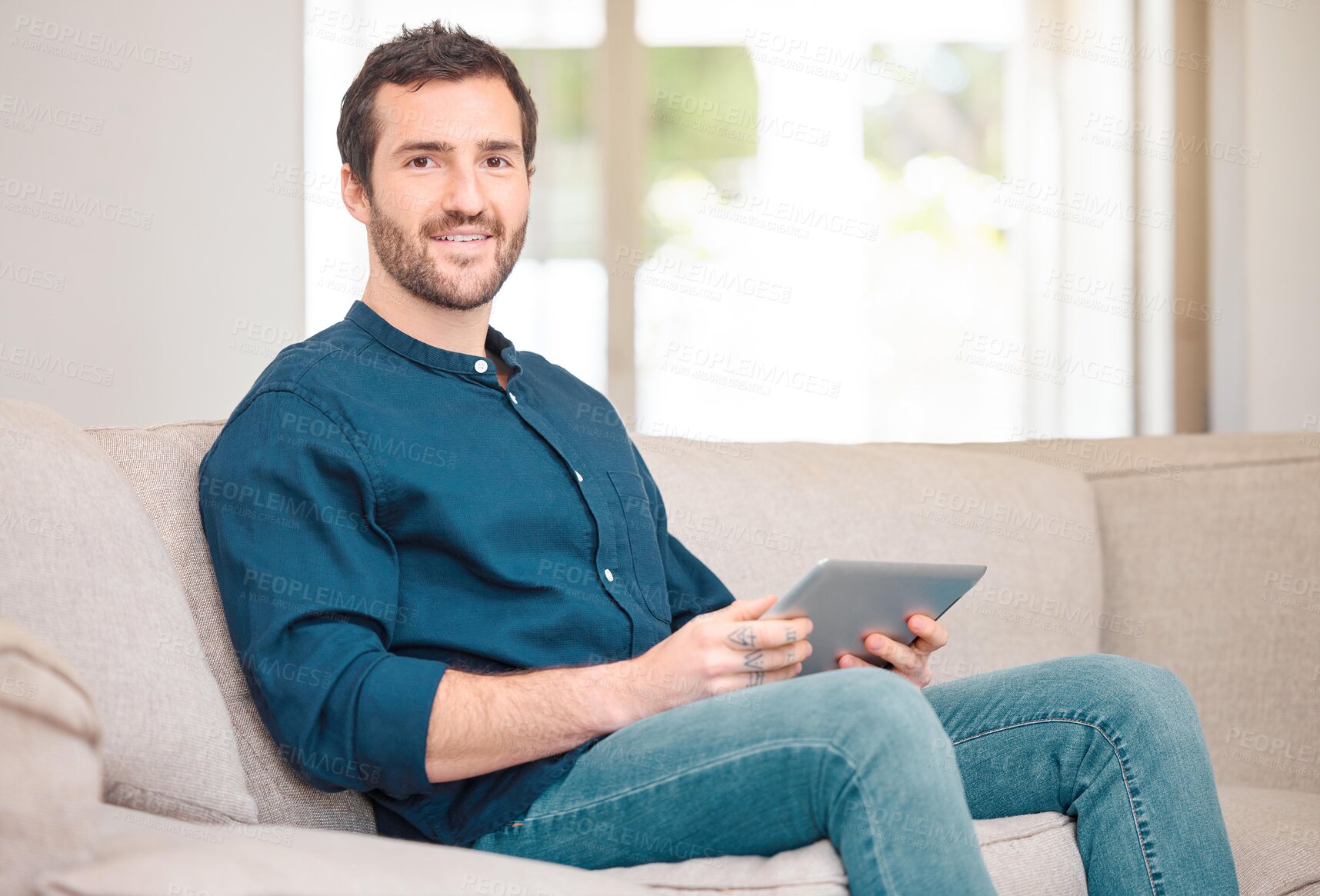 Buy stock photo Cropped portrait of a handsome young man using his tablet while sitting on the sofa at home